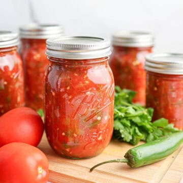 Jars of salsa for canning are arranged on a wooden board with fresh tomatoes, a green chili pepper, and cilantro in the foreground.