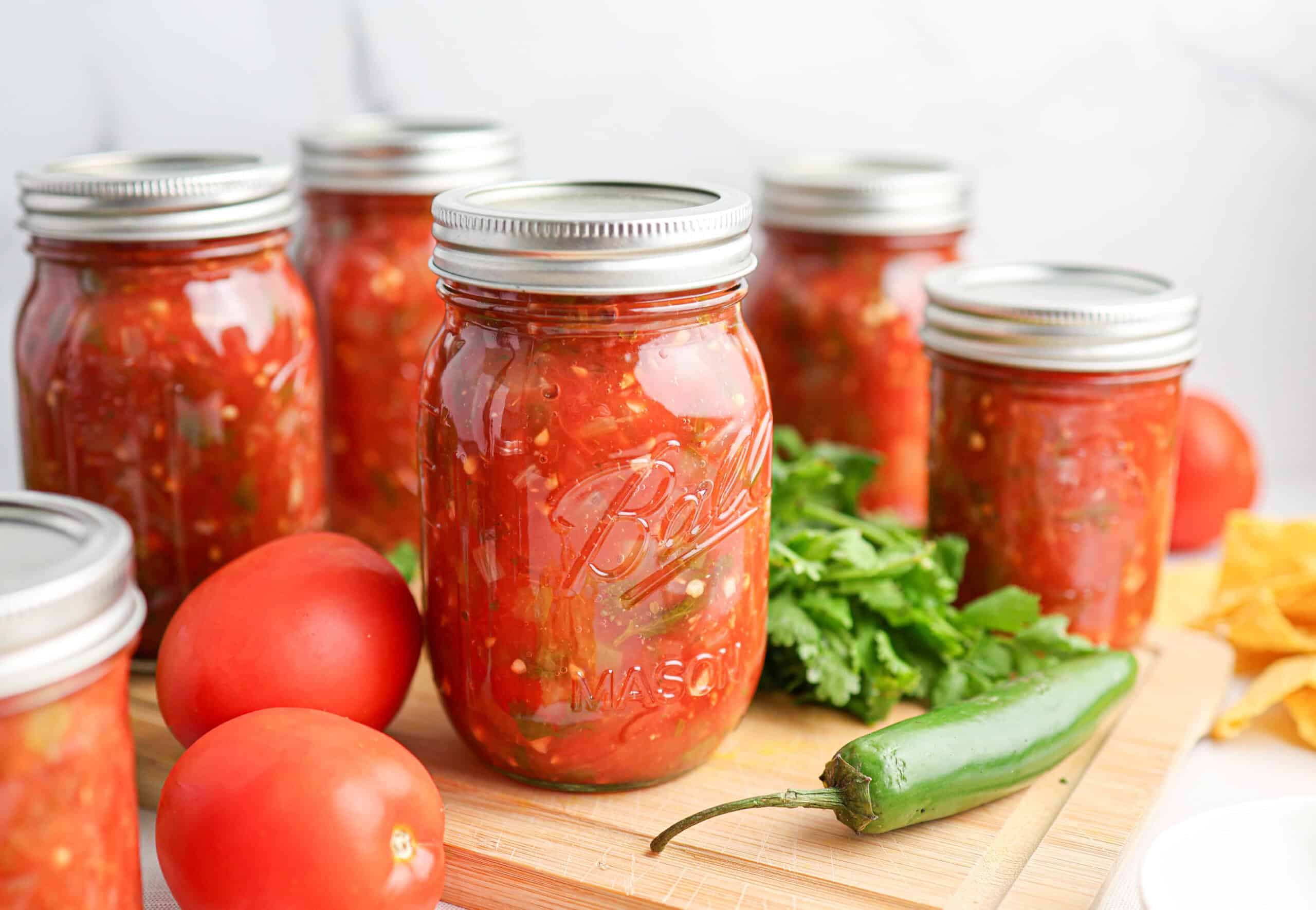 Jars of salsa for canning are arranged on a wooden board with fresh tomatoes, a green chili pepper, and cilantro in the foreground.
