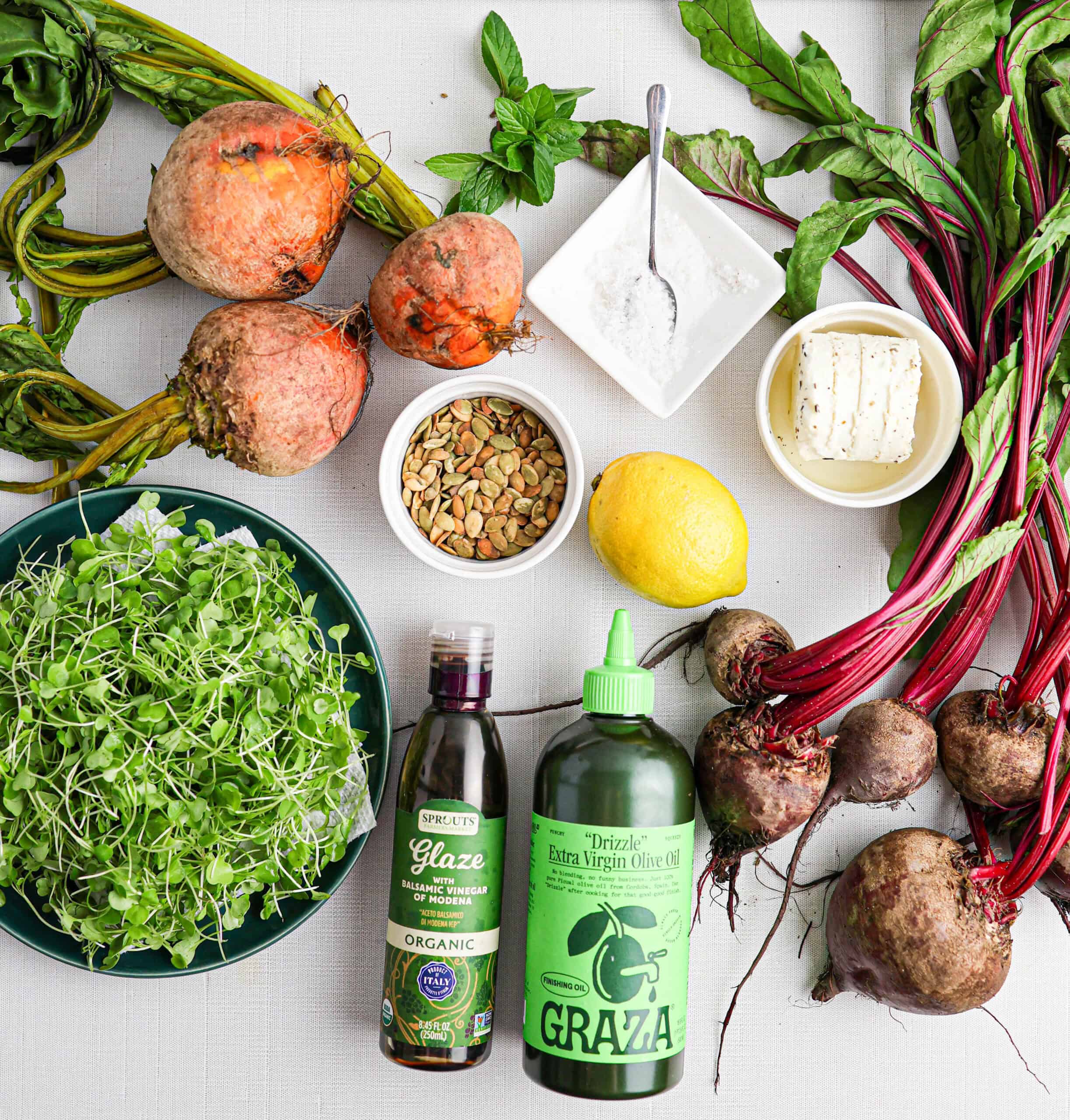 Assorted fresh beetroot with greens, lemon, feta, microgreens, fresh herbs, nuts, olive oil, balsamic glaze, and a small bowl of salt arranged on a white surface create a vibrant and delicious beet salad.