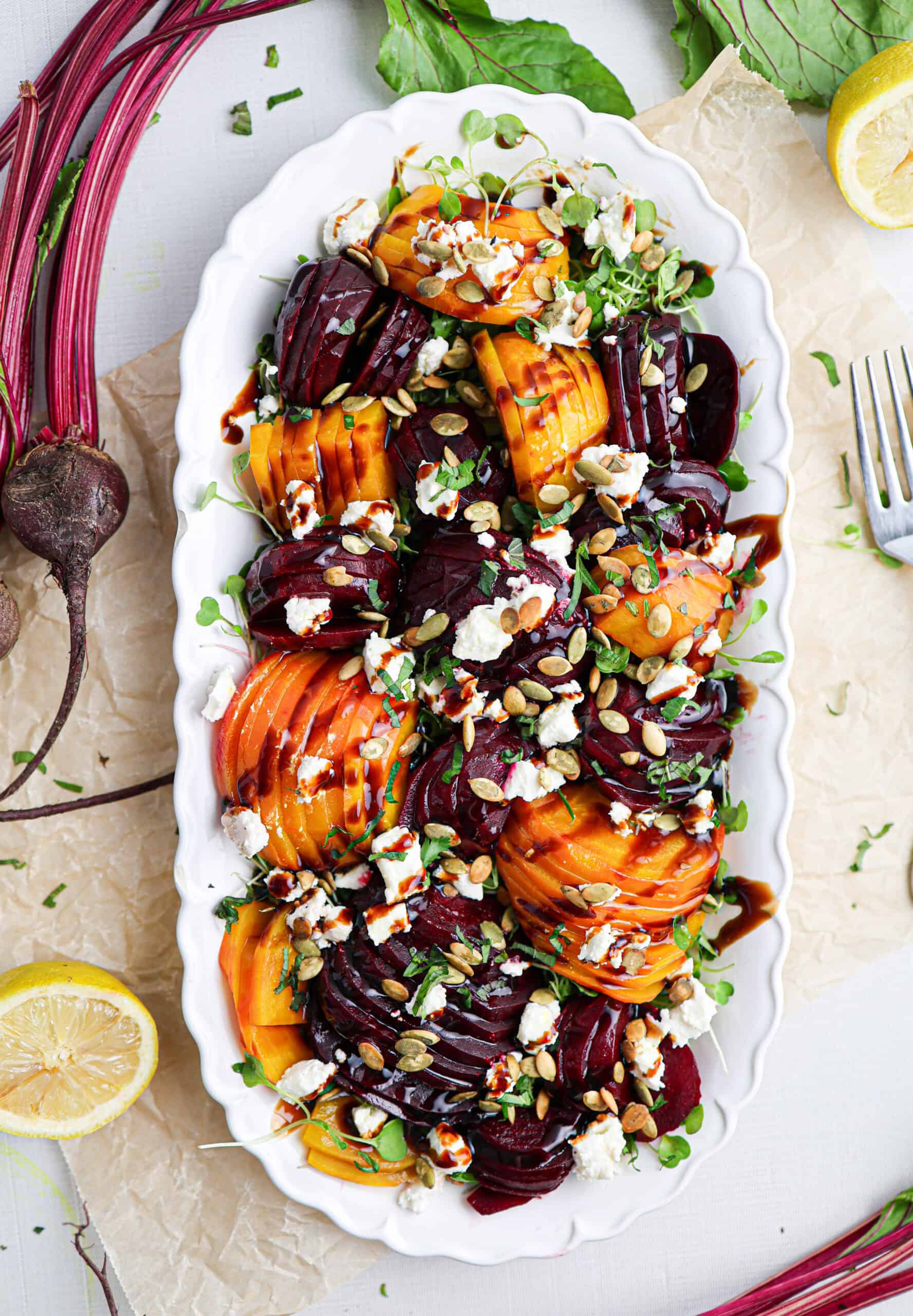 A white platter with beet salad featuring sliced red and golden beets, crumbled cheese, pumpkin seeds, microgreens, and balsamic drizzle, garnished with lemon slices.