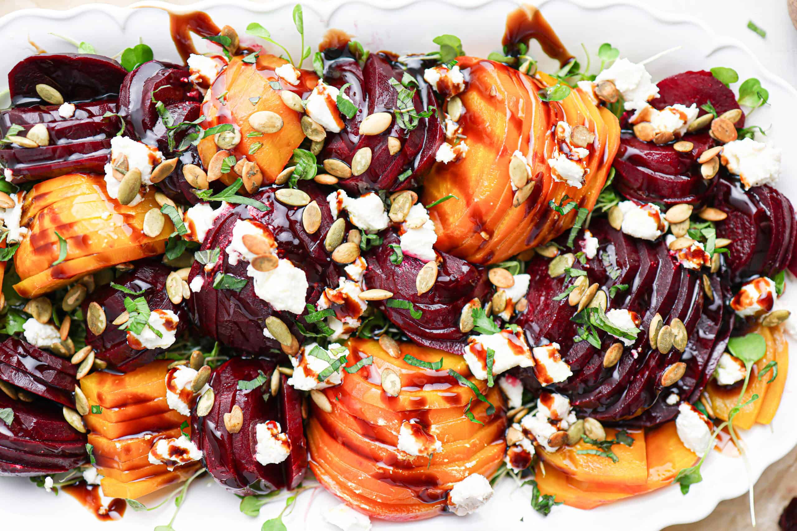 Sliced red and golden beets create a vibrant beet salad, topped with goat cheese, pumpkin seeds, microgreens, and a drizzle of balsamic glaze on a white platter.