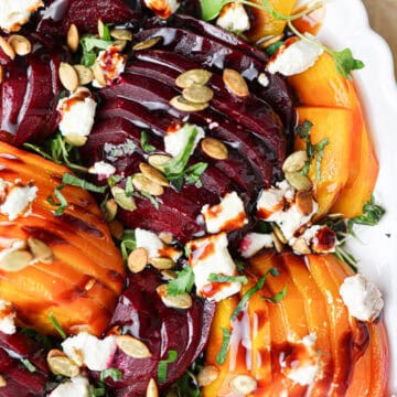 Sliced red and yellow beets on a white plate create a vibrant beet salad, topped with crumbled cheese, pumpkin seeds, fresh herbs, and a drizzle of balsamic glaze.