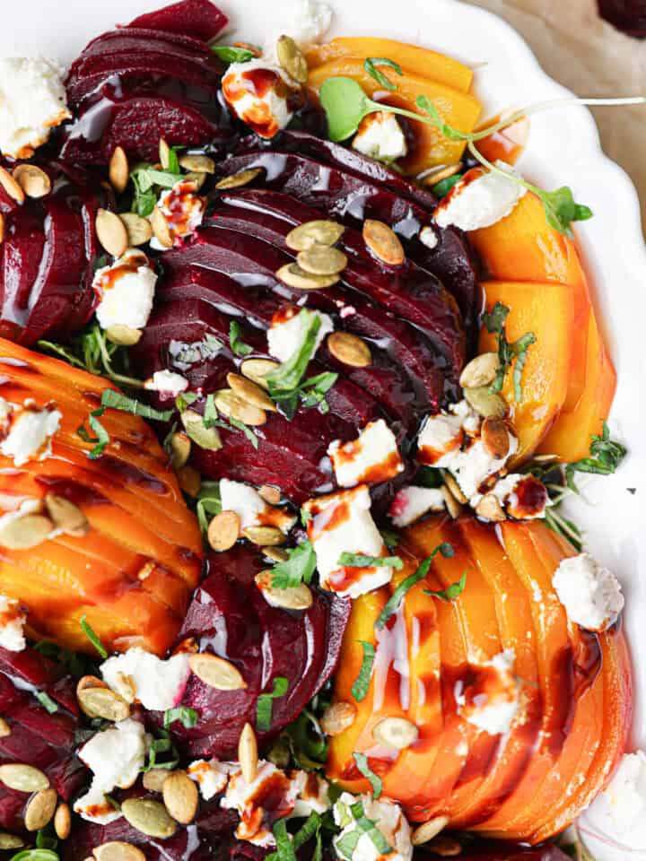 Sliced red and yellow beets on a white plate create a vibrant beet salad, topped with crumbled cheese, pumpkin seeds, fresh herbs, and a drizzle of balsamic glaze.