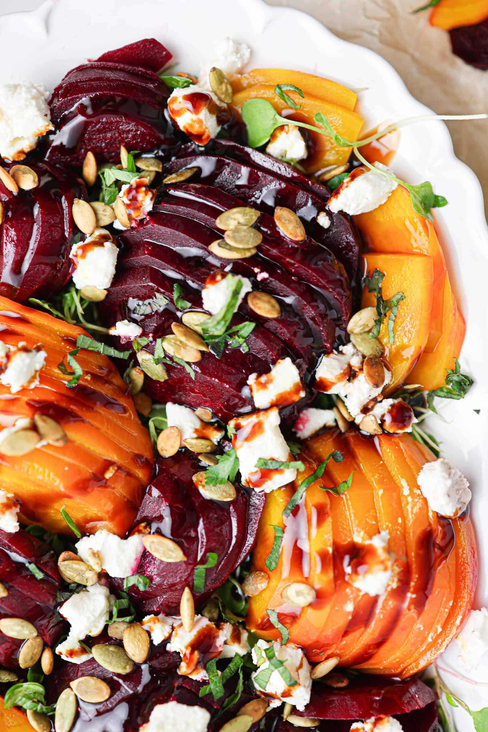 Sliced red and yellow beets on a white plate create a vibrant beet salad, topped with crumbled cheese, pumpkin seeds, fresh herbs, and a drizzle of balsamic glaze.