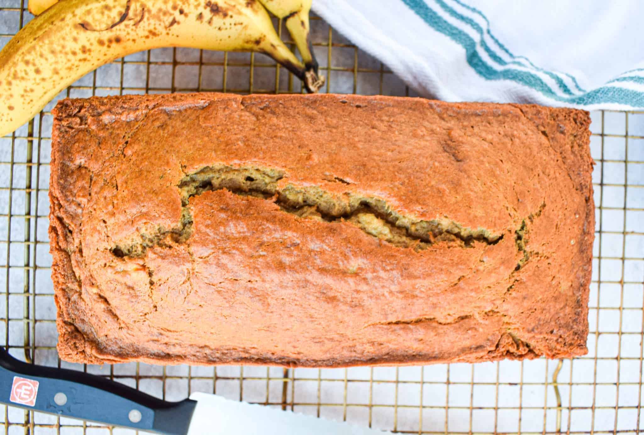 A loaf of the best easy banana bread with a crack down the center rests on a cooling rack beside a knife and two ripe bananas.