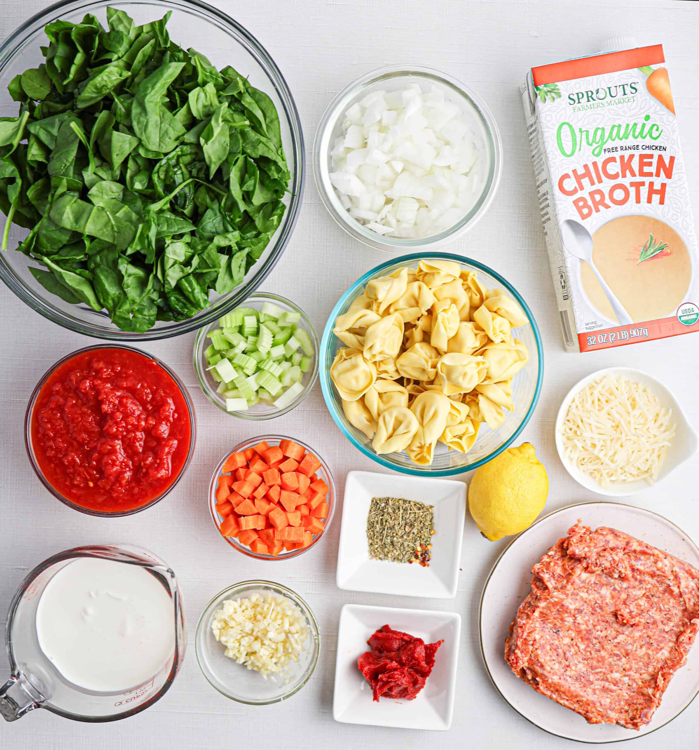 Ingredients for tortellini soup are artfully arranged on a white surface, featuring spinach, onions, chicken broth, tortellini, ground meat, tomatoes, carrots, celery, garlic, cream, tomato paste, cheese, and an array of spices.
