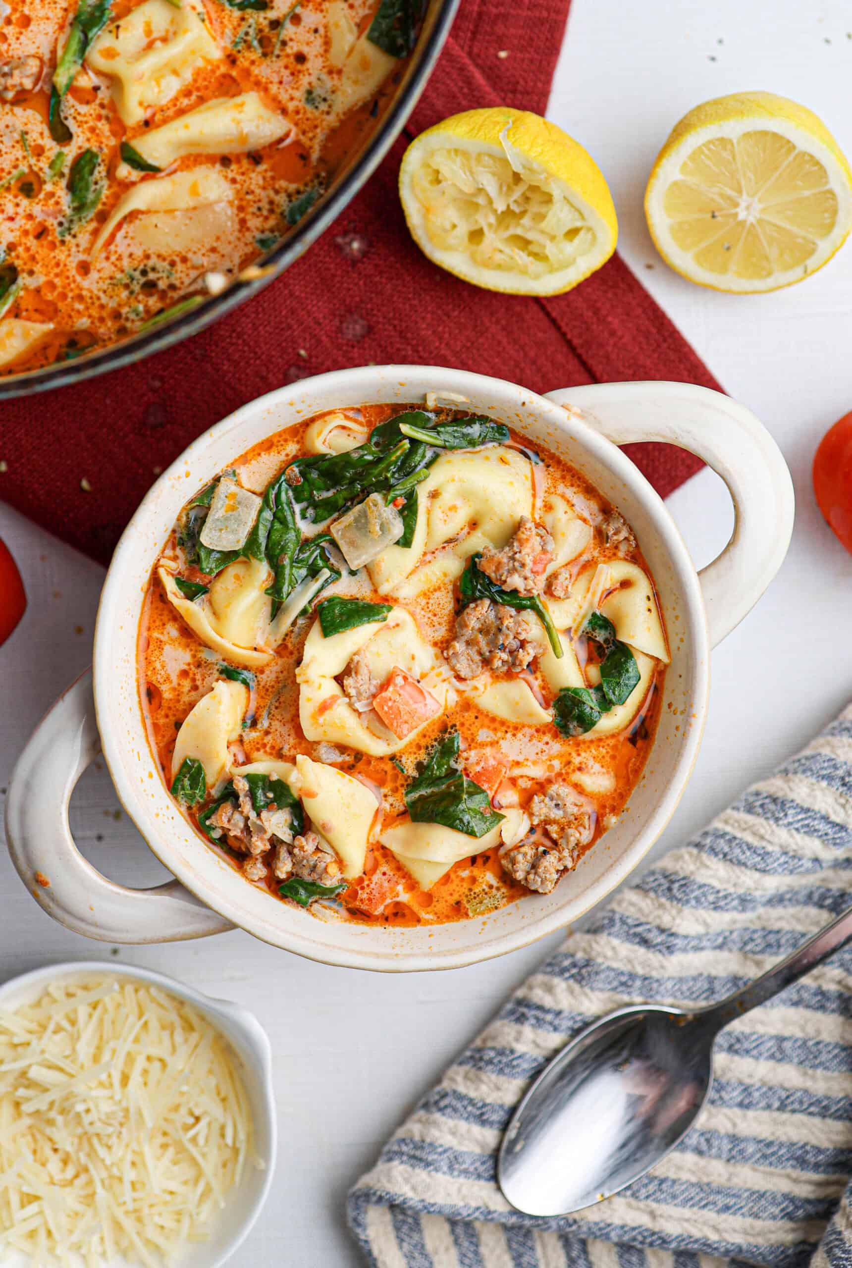 A bowl of creamy tortellini soup with ground meat, spinach, and onions, surrounded by lemon halves, shredded cheese, and a spoon on a striped cloth. This delicious tortellini soup is both comforting and satisfying—a perfect meal for any occasion.