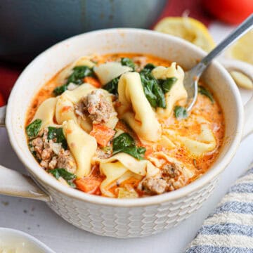 A bowl of creamy tortellini soup with ground meat, spinach, and carrots, with a spoon resting inside the bowl.