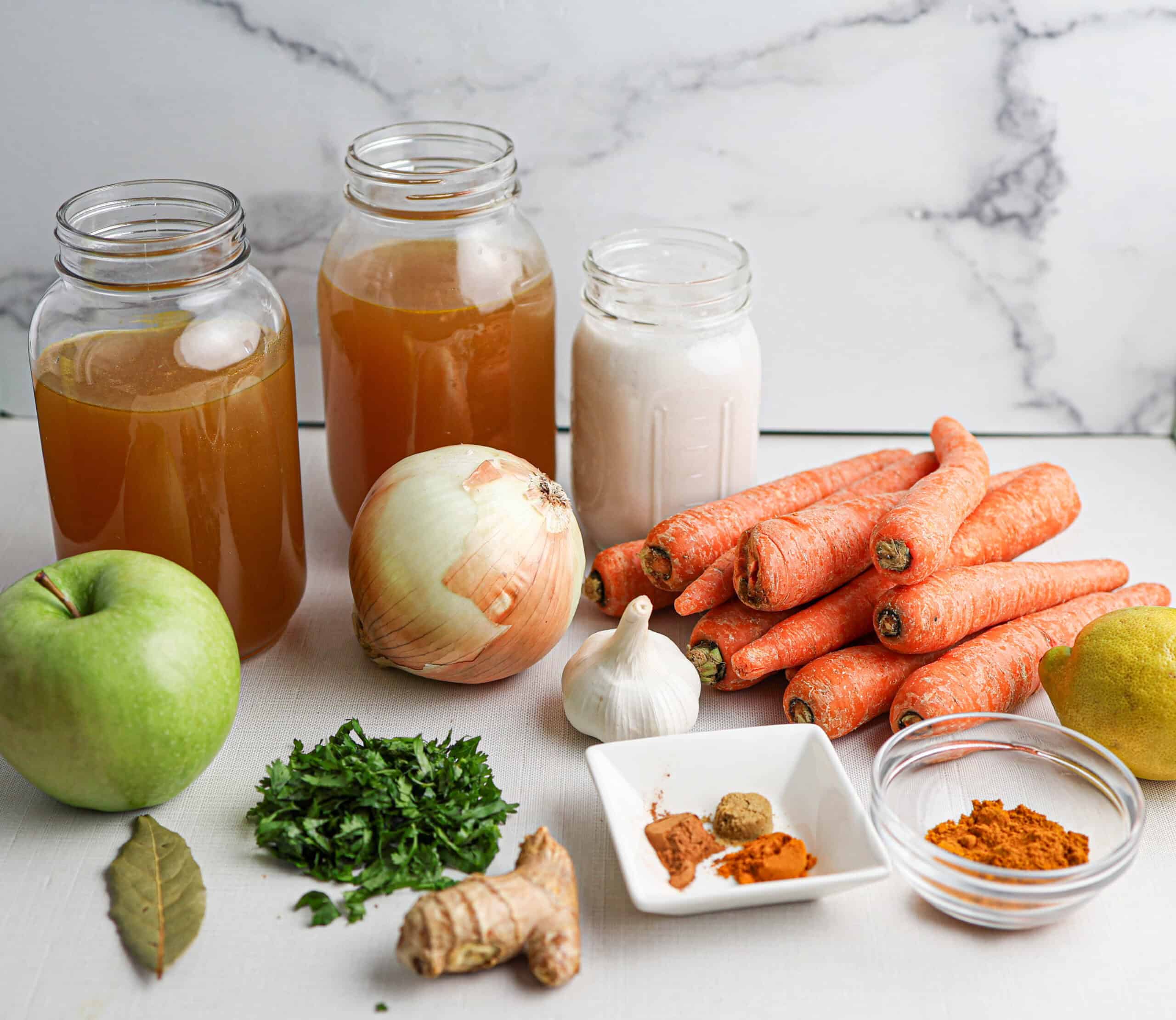 Various ingredients are arranged on a counter for making carrot soup, including carrots, onion, apple, garlic, fresh herbs, ginger, spices, bay leaf, two jars of broth, and a jar of a light-colored liquid.