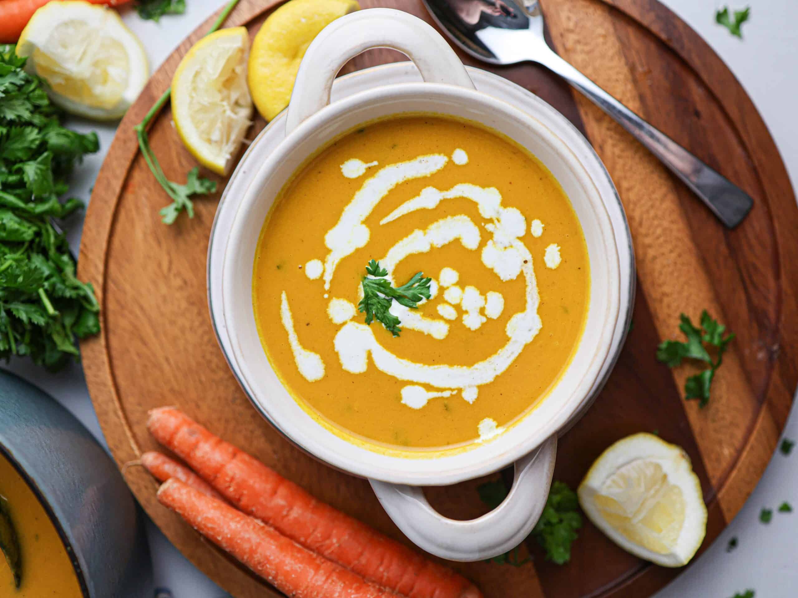 A bowl of creamy carrot soup, its vibrant orange hue garnished with a swirl of cream and parsley, is surrounded by fresh carrots, lemon slices, and fragrant herbs on a wooden board.