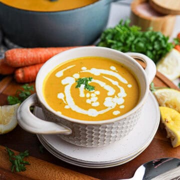 A bowl of creamy orange carrot soup garnished with cream and parsley, surrounded by fresh carrots, herbs, lemon, and a pot of soup in the background.