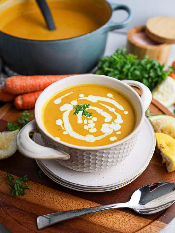 A bowl of creamy orange carrot soup garnished with cream and parsley, surrounded by fresh carrots, herbs, lemon, and a pot of soup in the background.