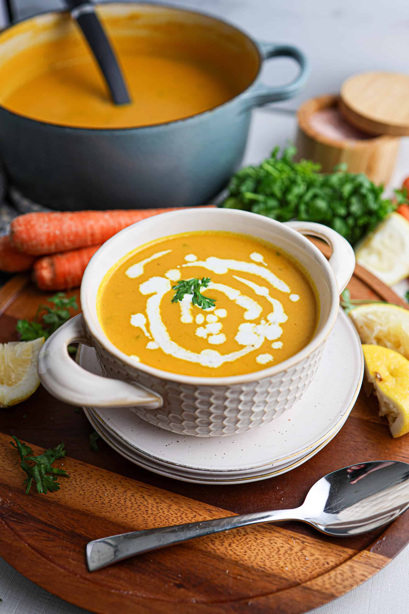 A bowl of creamy orange carrot soup garnished with cream and parsley, surrounded by fresh carrots, herbs, lemon, and a pot of soup in the background.