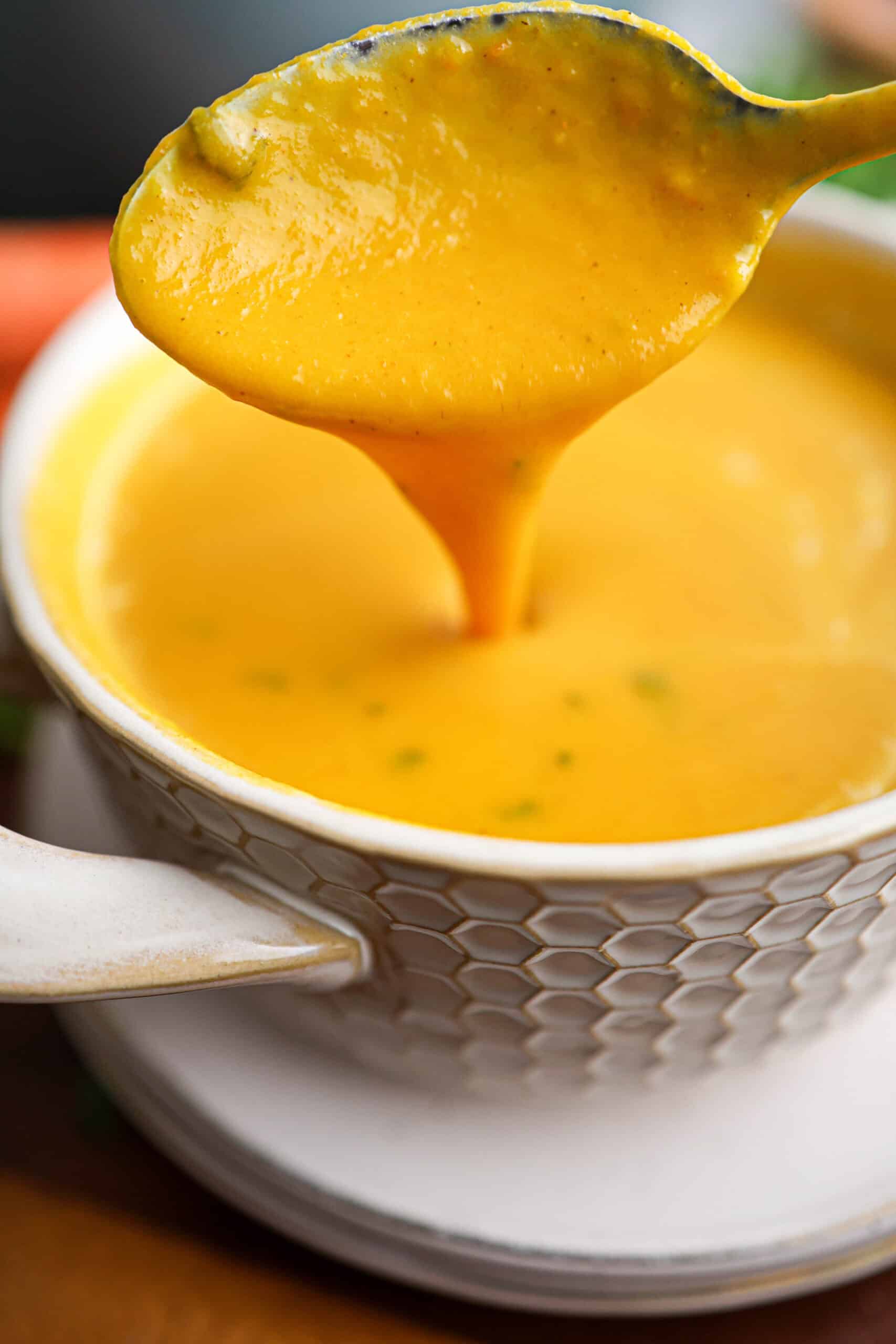 A spoon lifts a portion of creamy carrot soup from a white textured bowl sitting on a saucer.