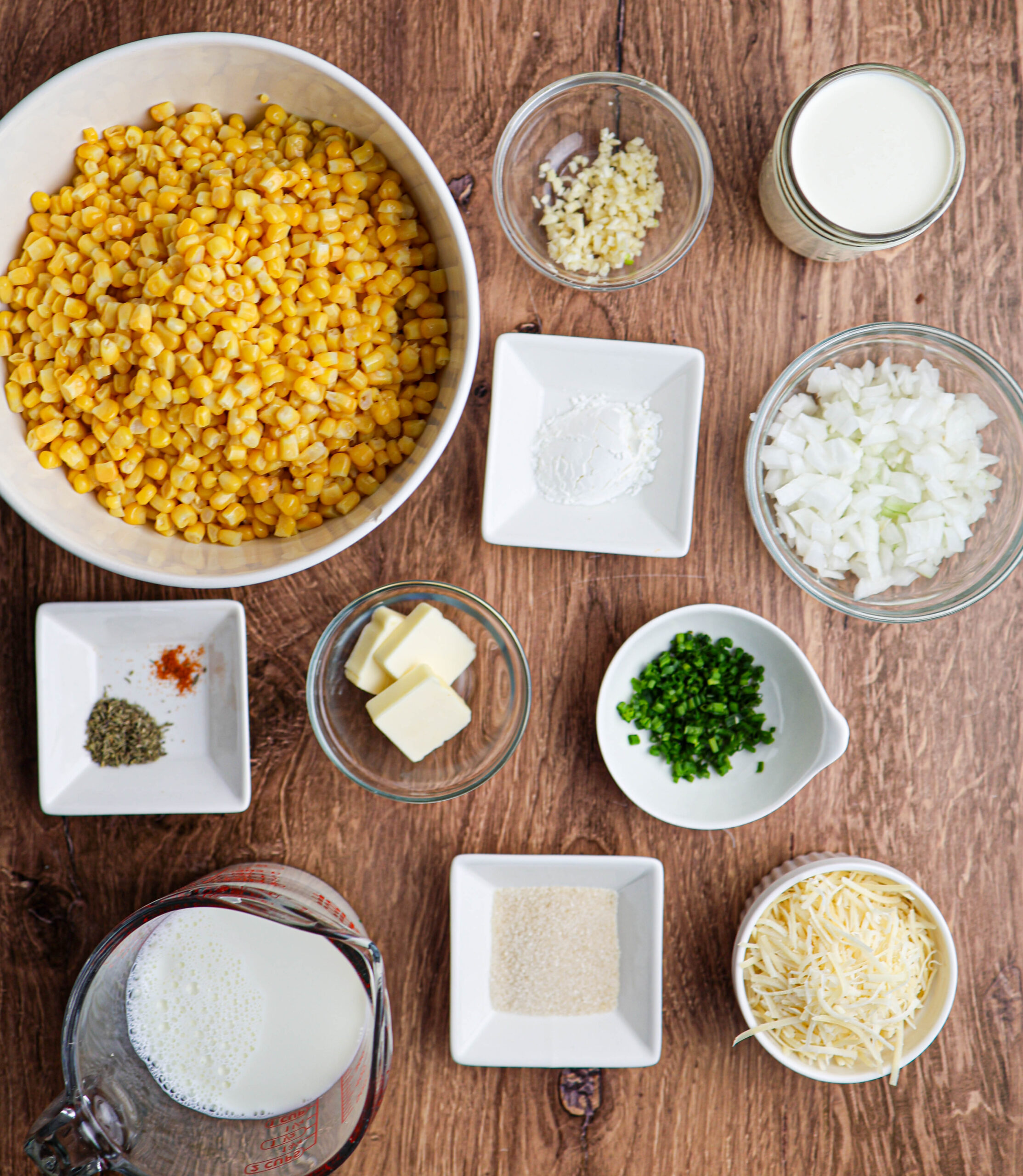 Ingredients for creamed corn are artfully arranged on a wooden surface, including corn, onion, garlic, milk, butter, cheese, flour, chopped jalapeño, spices, and sugar in separate bowls and dishes.