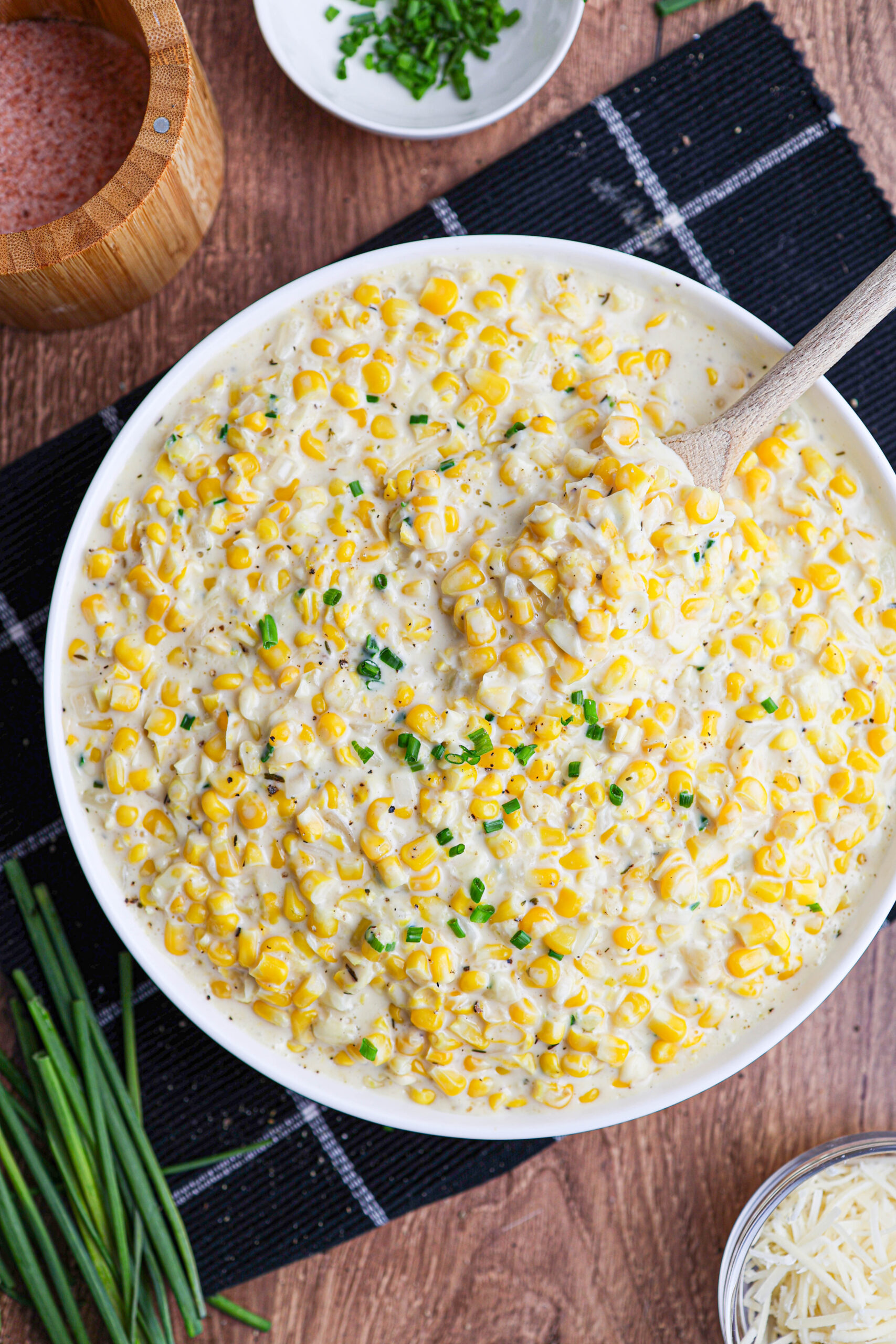 A bowl of creamy corn casserole made with creamed corn and garnished with chopped chives sits on a black cloth, with a wooden spoon, wooden salt container, and extra chives nearby.