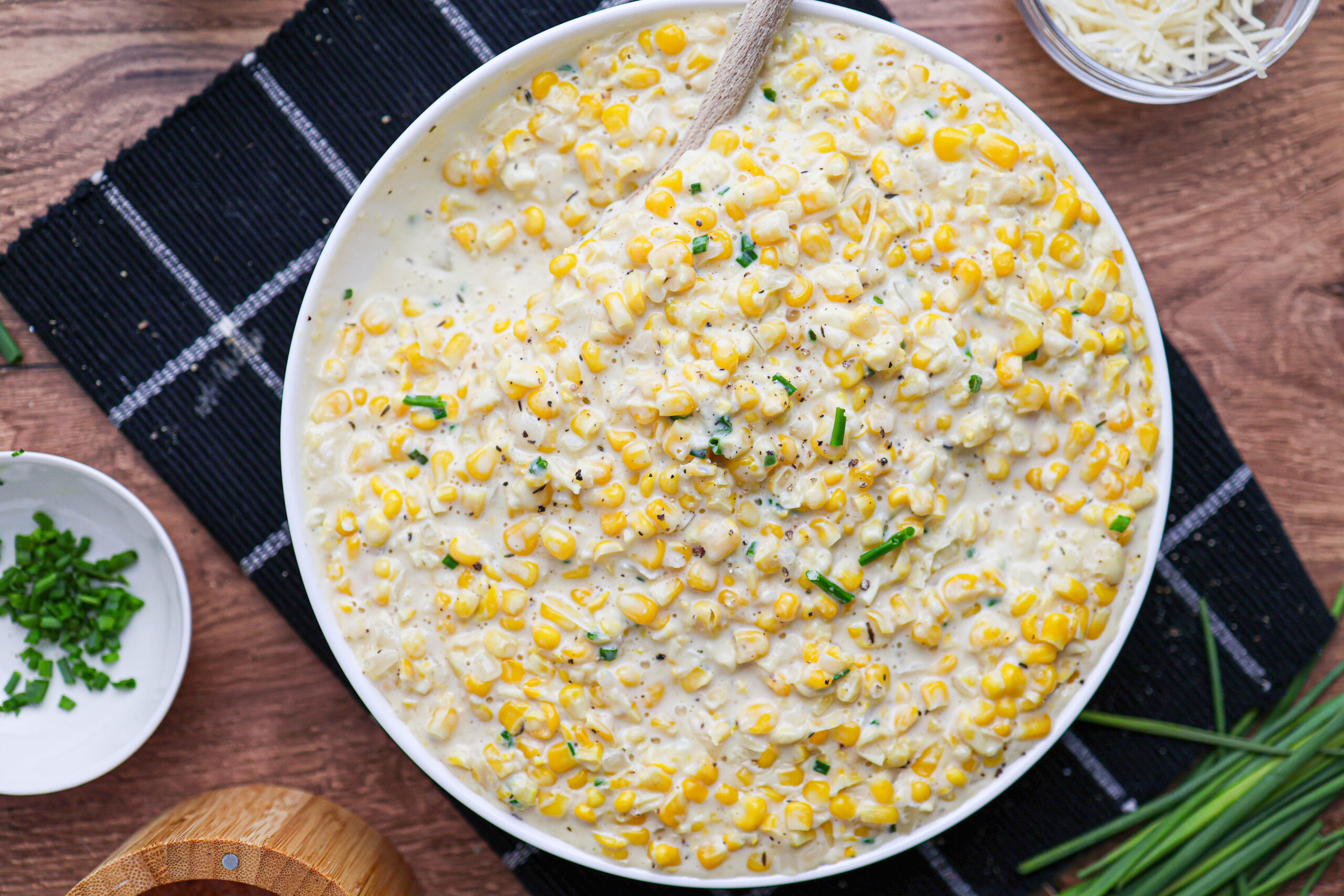 A bowl filled with creamy creamed corn topped with chopped chives, with a spoon inside, sits on a black placemat on a wooden table.