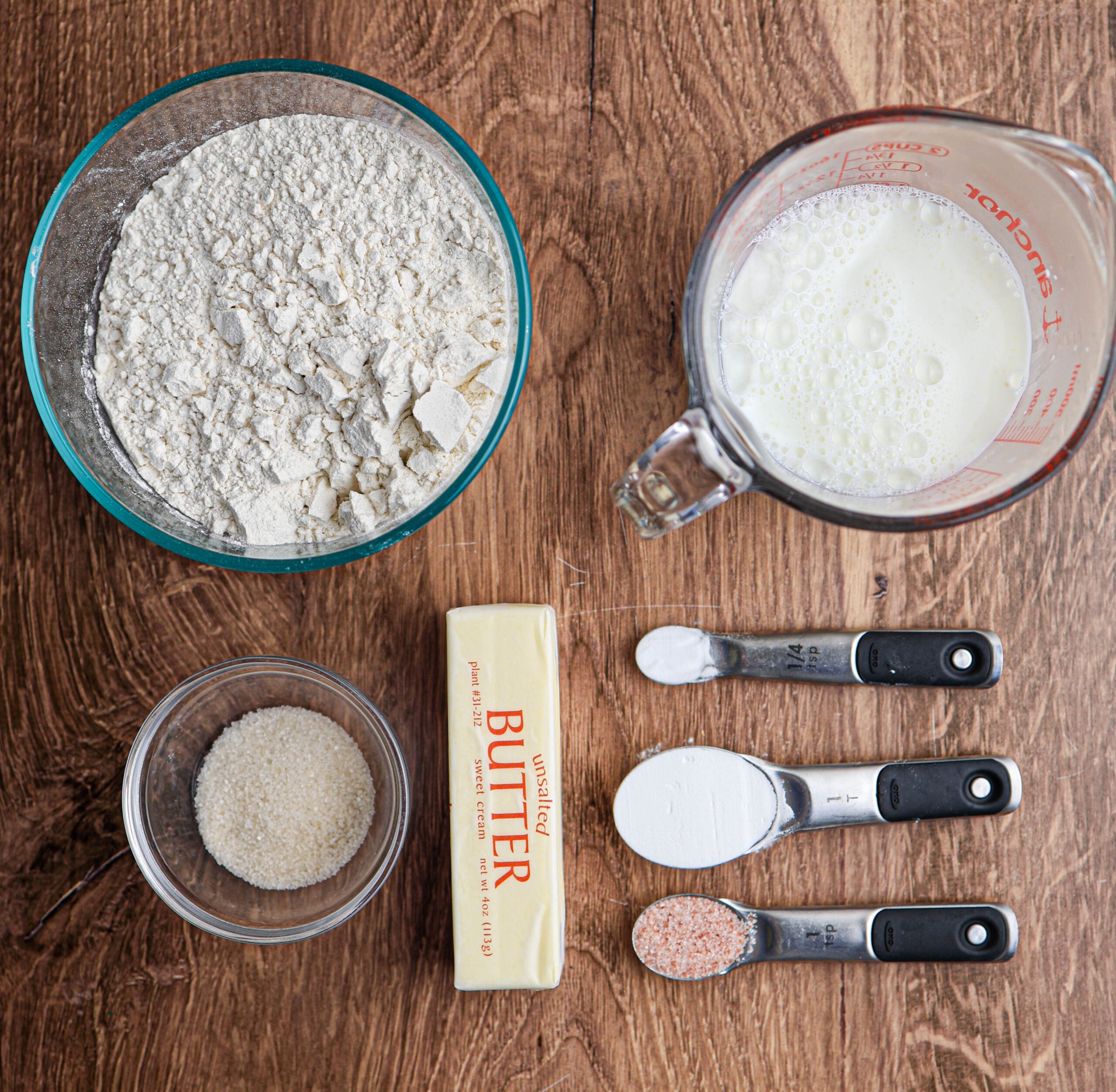 Flour, milk, sugar, butter, baking powder, salt, and a measuring spoon of flour are arranged on a wooden surface—perfect ingredients for making classic buttermilk biscuits.