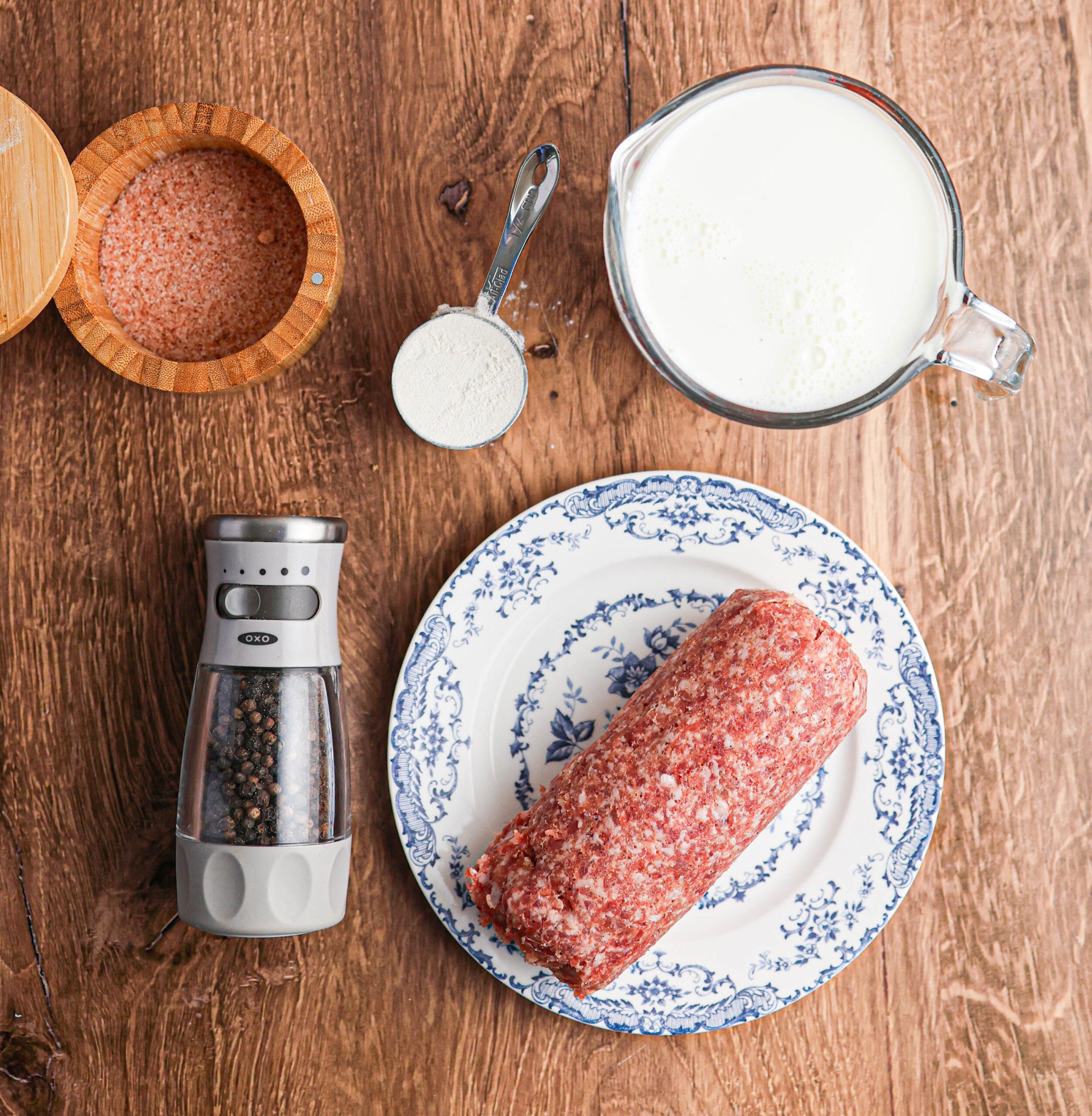 Ground meat on a plate, a measuring cup of milk, a small bowl of salt, a spoon of flour, and a pepper grinder on a wooden surface—all the essential ingredients for making delicious sausage gravy.