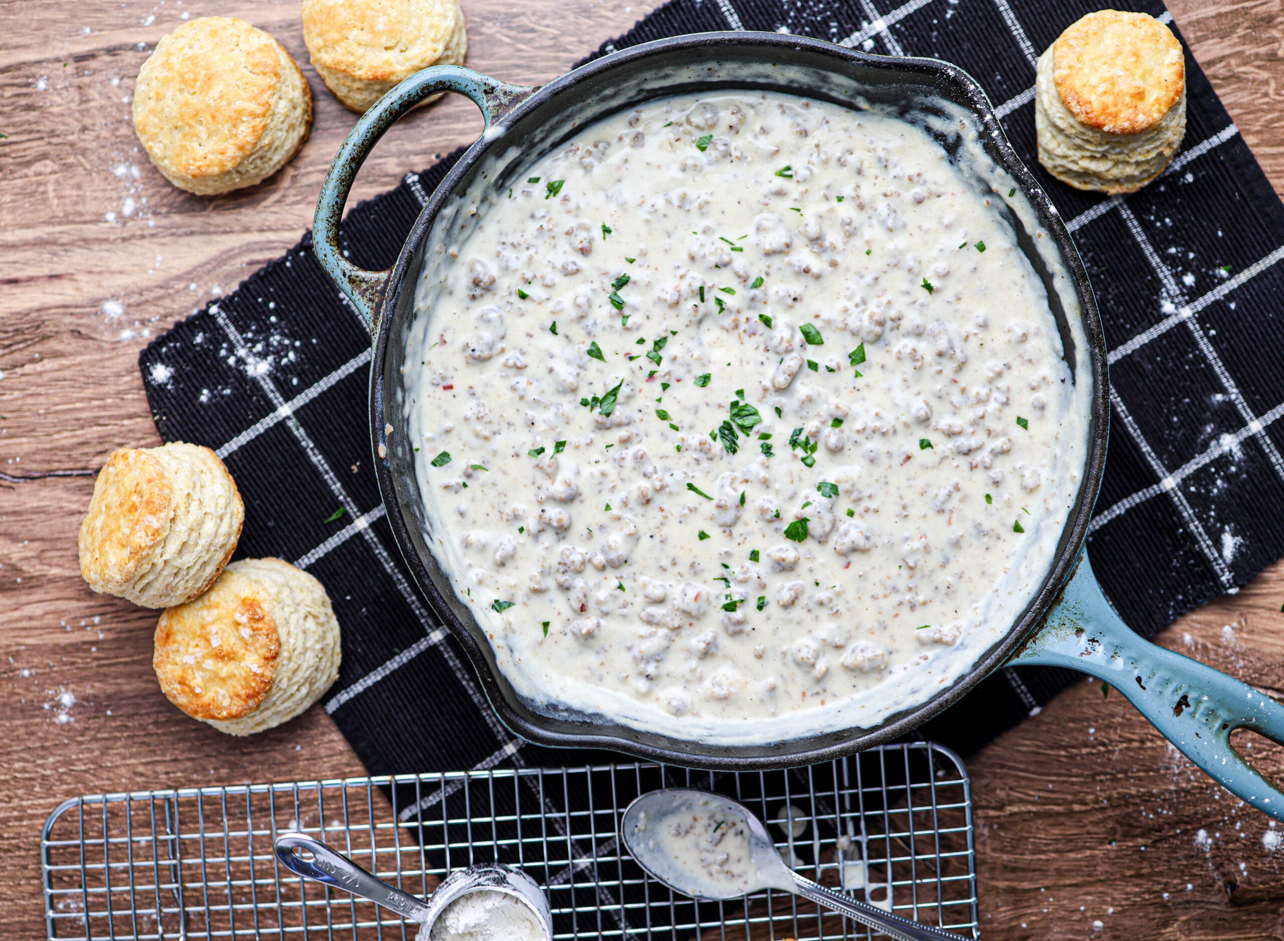 A skillet of savory sausage gravy garnished with herbs sits on a checkered cloth, surrounded by several golden biscuits, with a spoon and cooling rack nearby.