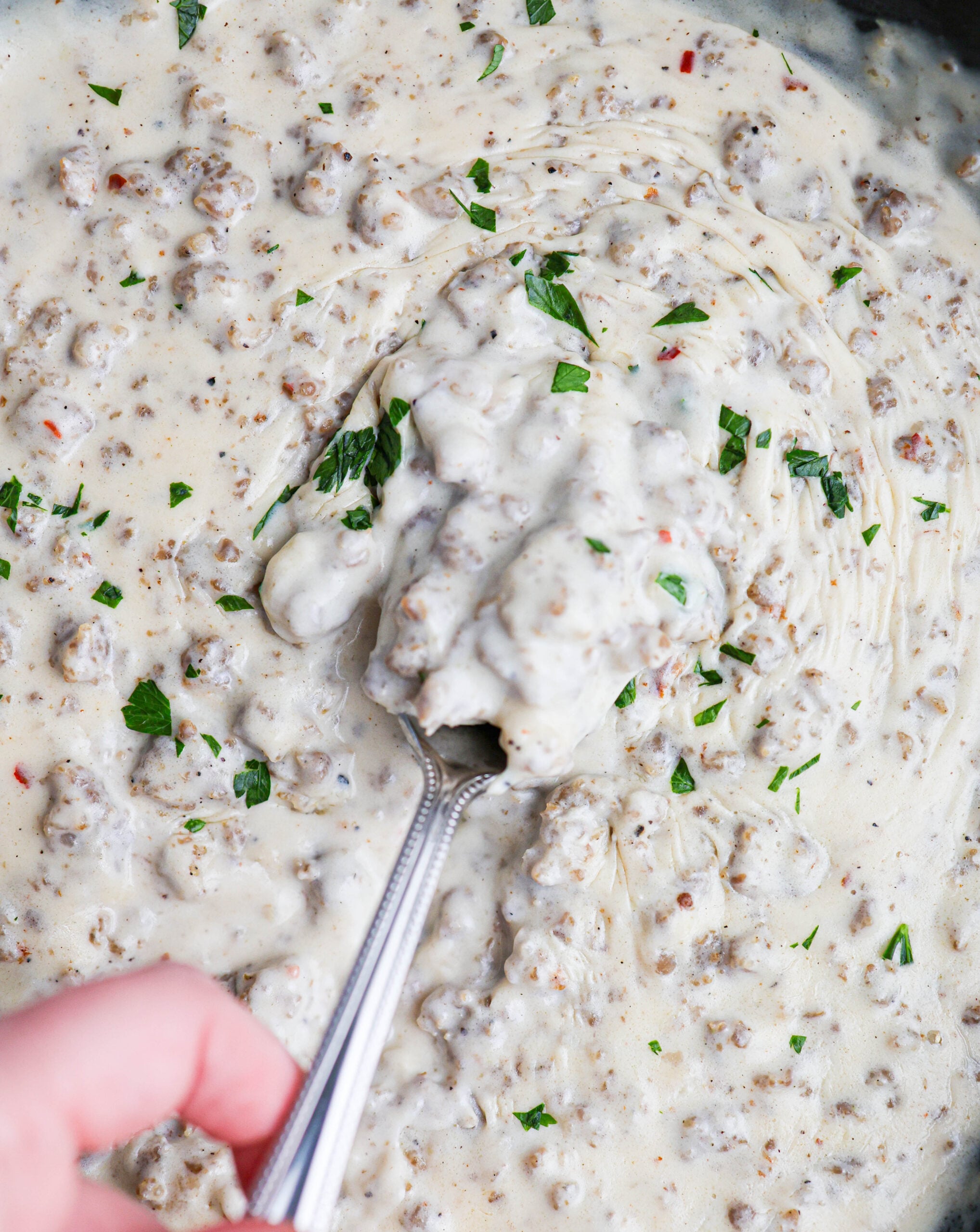 A hand holds a spoon, scooping rich and creamy sausage gravy with chopped parsley from a pan.