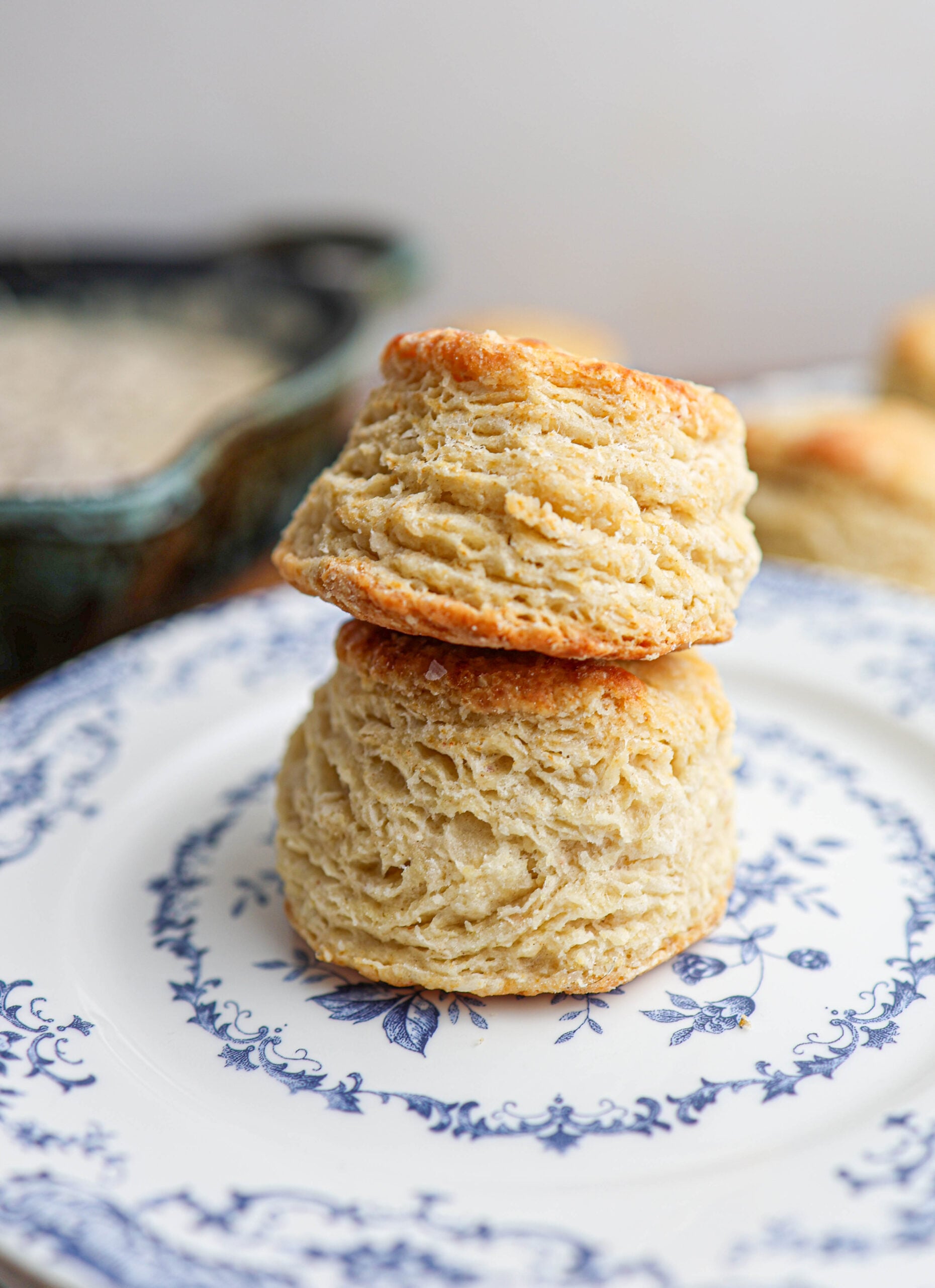 Two golden, flaky buttermilk biscuits are stacked on a white plate with blue floral patterns, with a blurred background of additional biscuits and a dish.