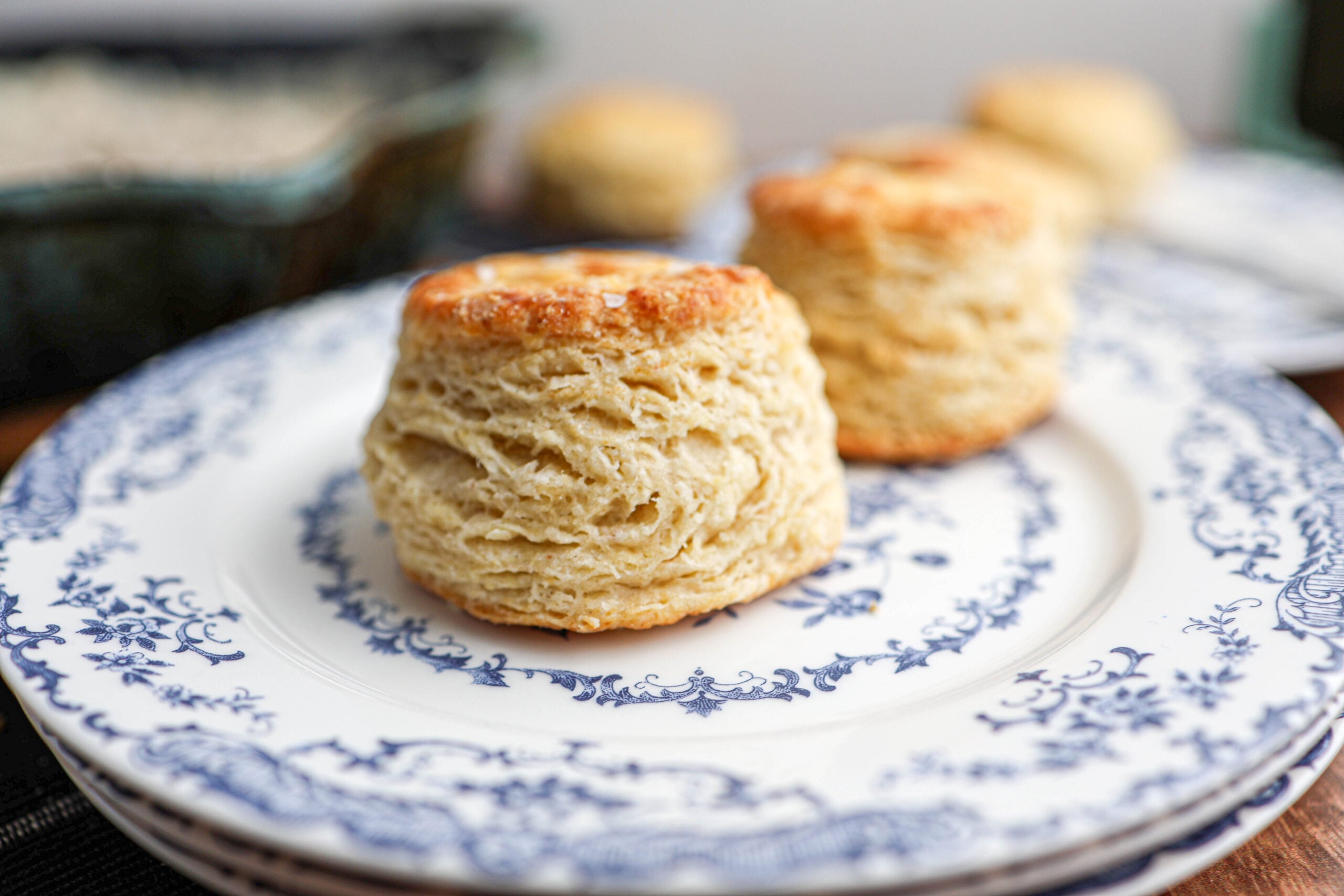 A golden-brown flaky biscuit sits on a white and blue patterned plate, with more biscuits visible in the background.