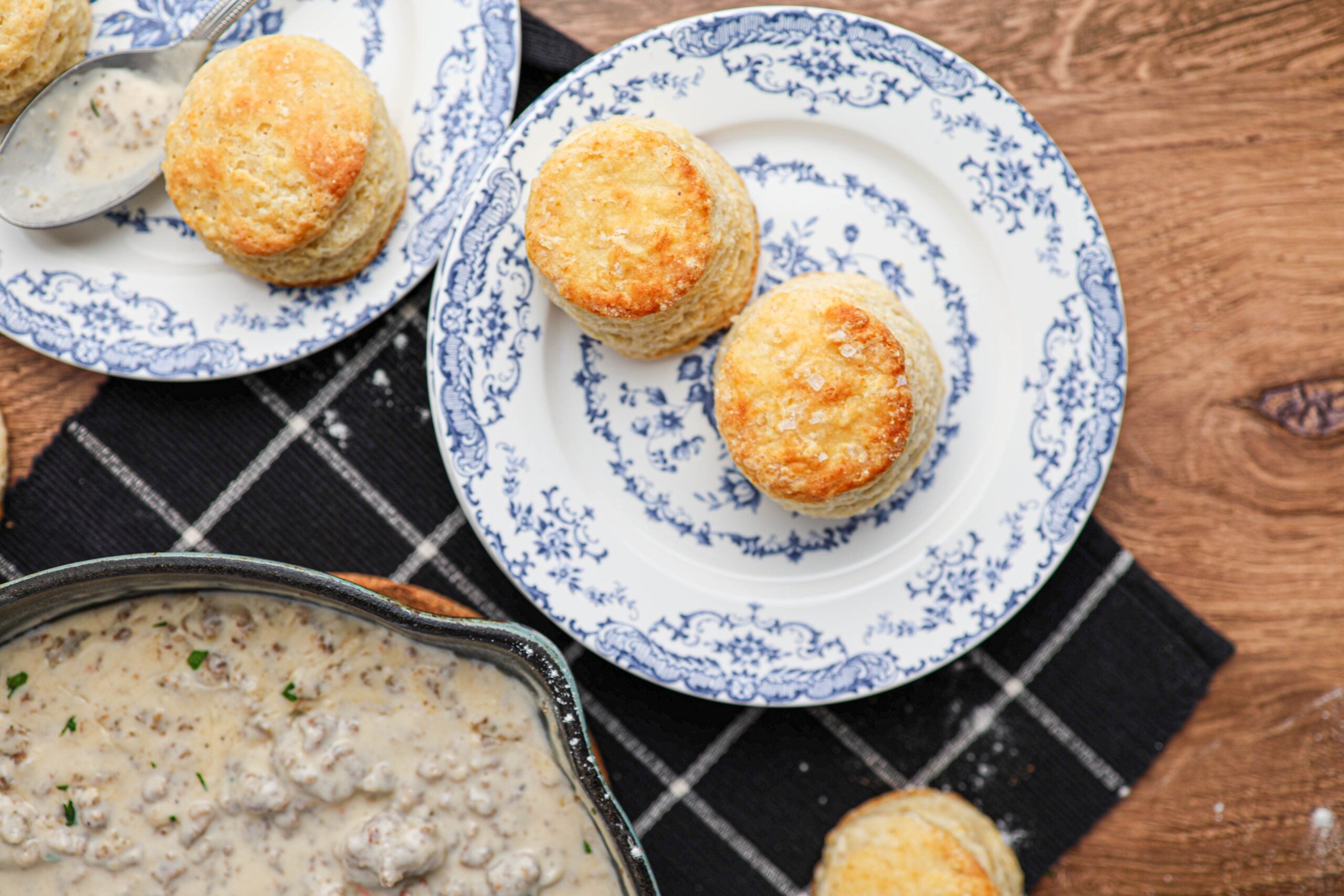 Buttermilk biscuits on blue and white plates sit next to a pan of sausage gravy, all arranged on a black checkered cloth over a wooden table.