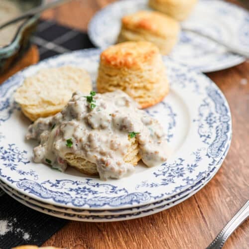 A biscuit smothered in savory sausage gravy is served on a patterned plate, with whole biscuits and a fork in the background.