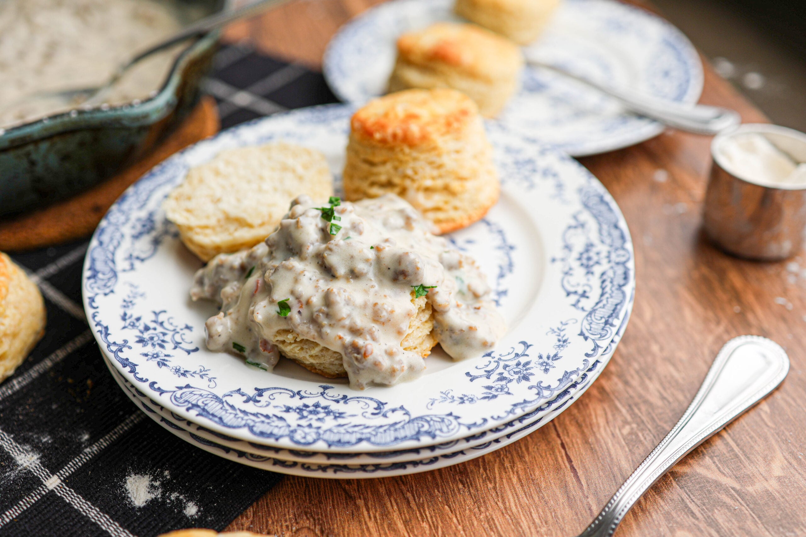 A generous serving of homemade biscuits and gravy served on a patterned plate, with whole flaky, buttermilk biscuits and a fork in the background.