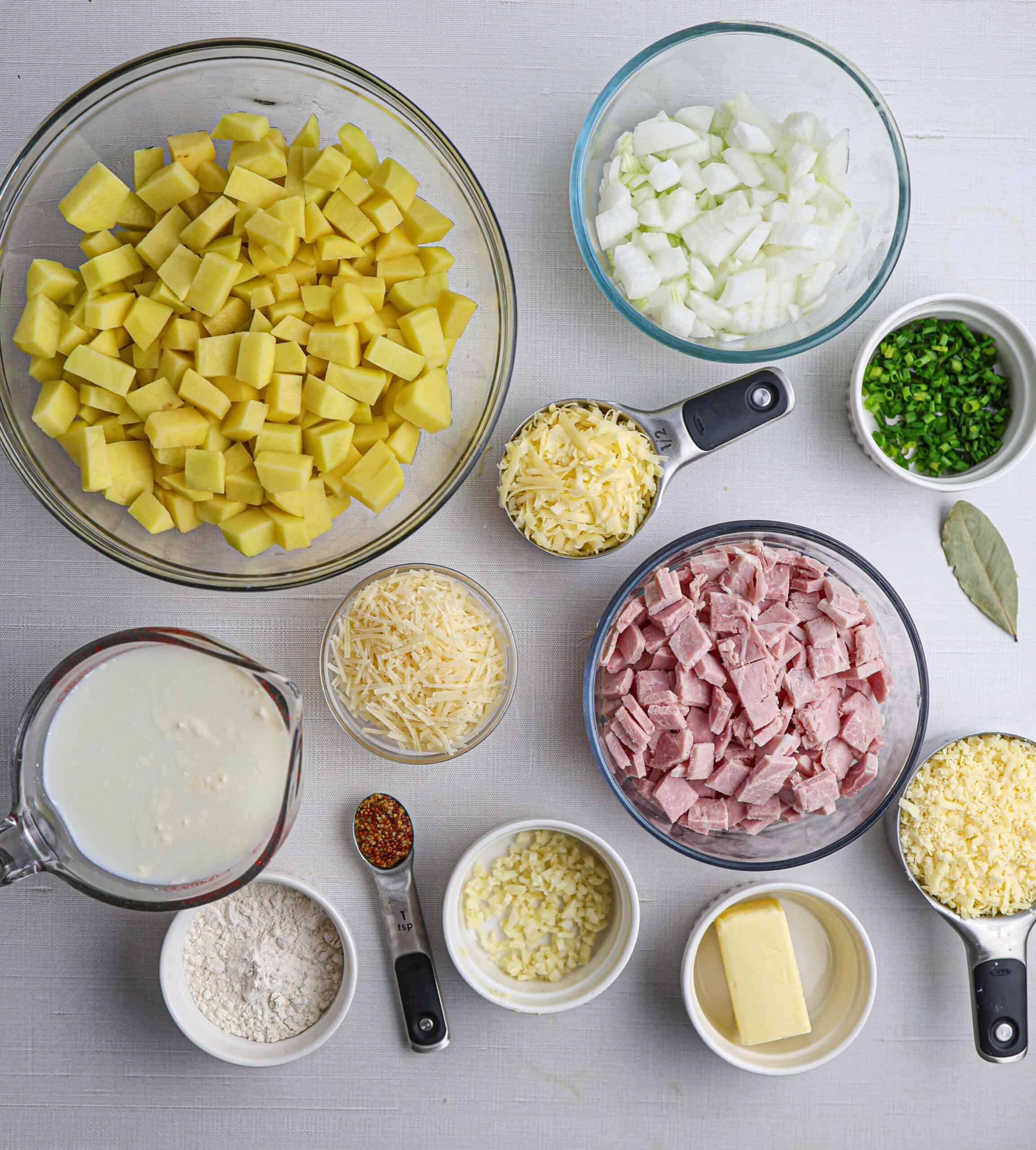 Ingredients for a ham and potato casserole, including diced potatoes, chopped onion, ham, shredded cheese, milk, flour, butter, garlic, a bay leaf, and a spoonful of mustard.