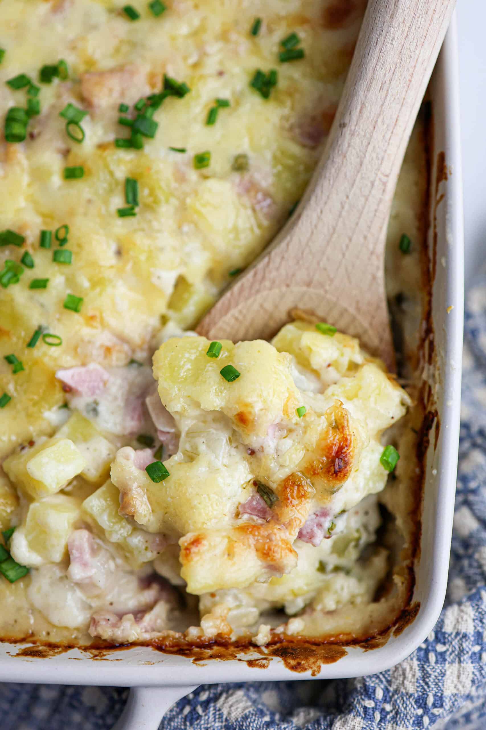 A wooden spoon lifting a serving of cheesy ham and potato casserole from a baking dish, garnished with chopped chives.