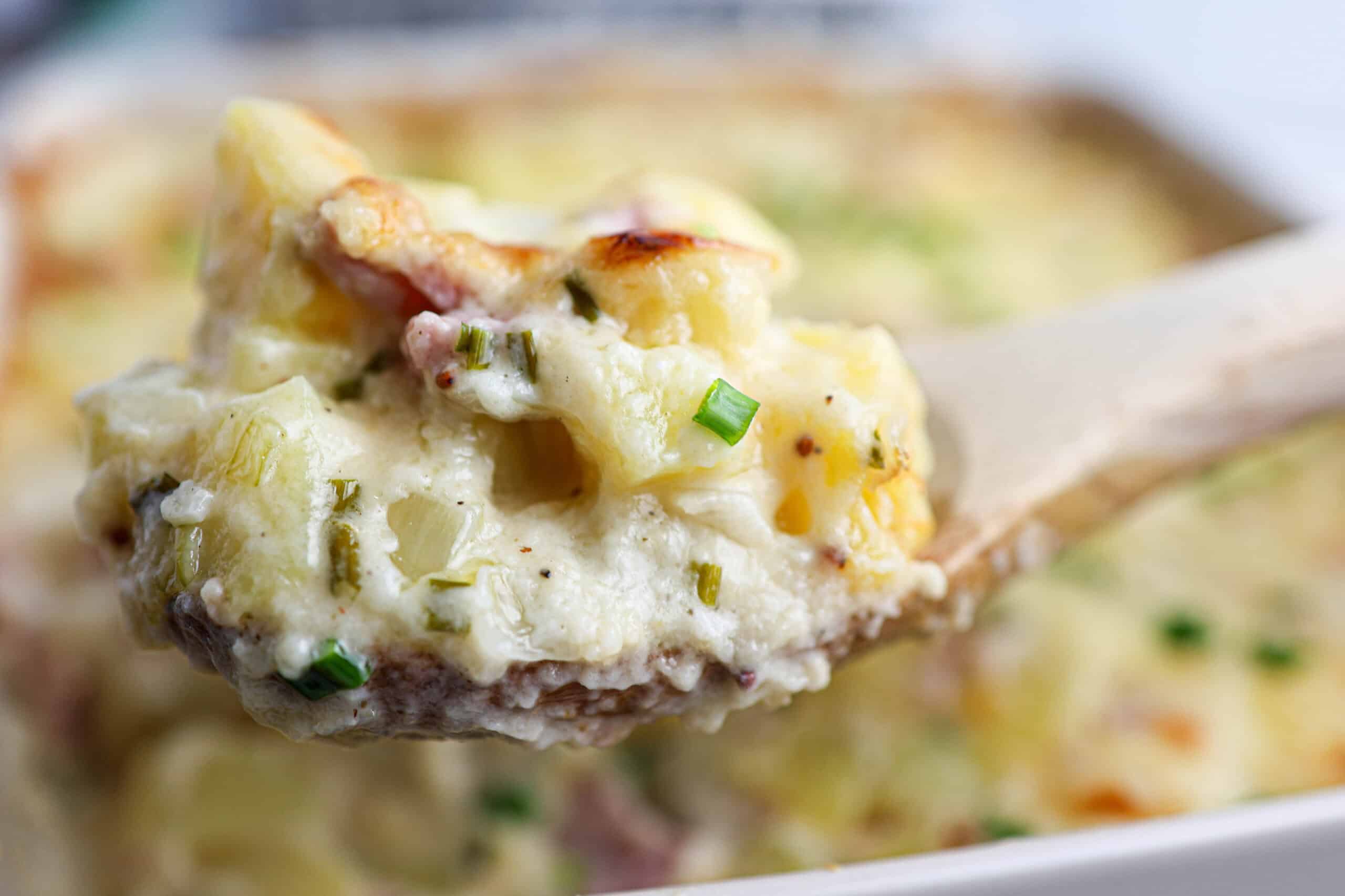 A wooden spoon holds a serving of creamy ham and potato casserole topped with chives.